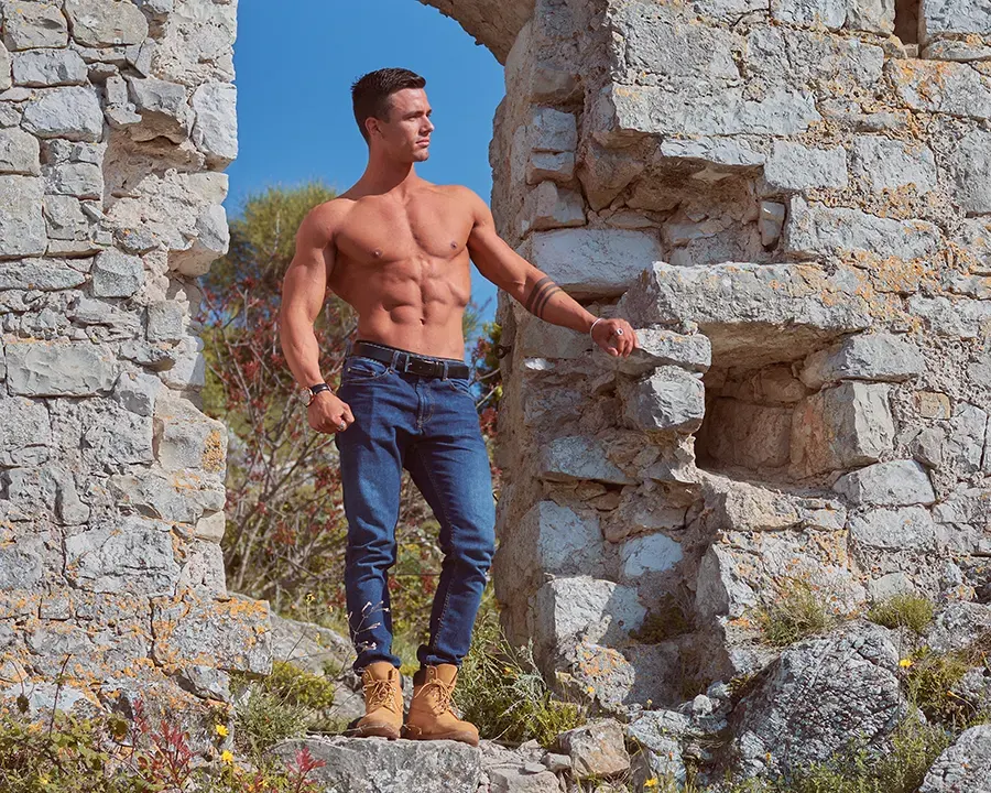 Athlète torse nu et musclé en jean posant dans des ruines de pierre au soleil, séance photo fitness en extérieur