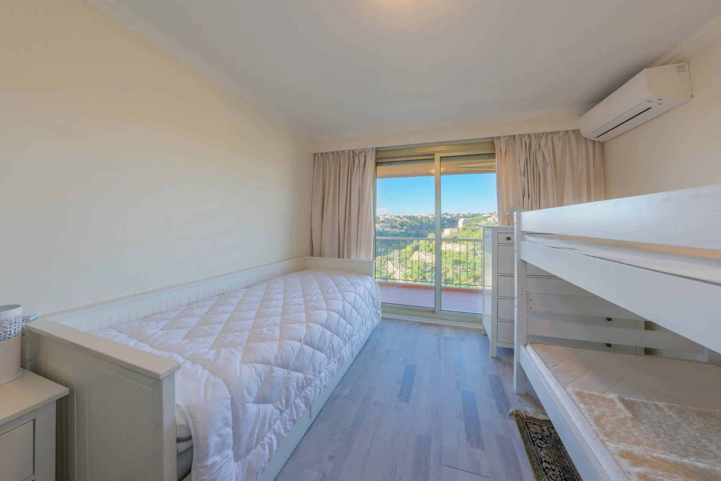 Chambre lumineuse avec lit gigogne blanc, lits superposés et baie vitrée sur balcon et collines, photographie immobilière en Suisse romande & France