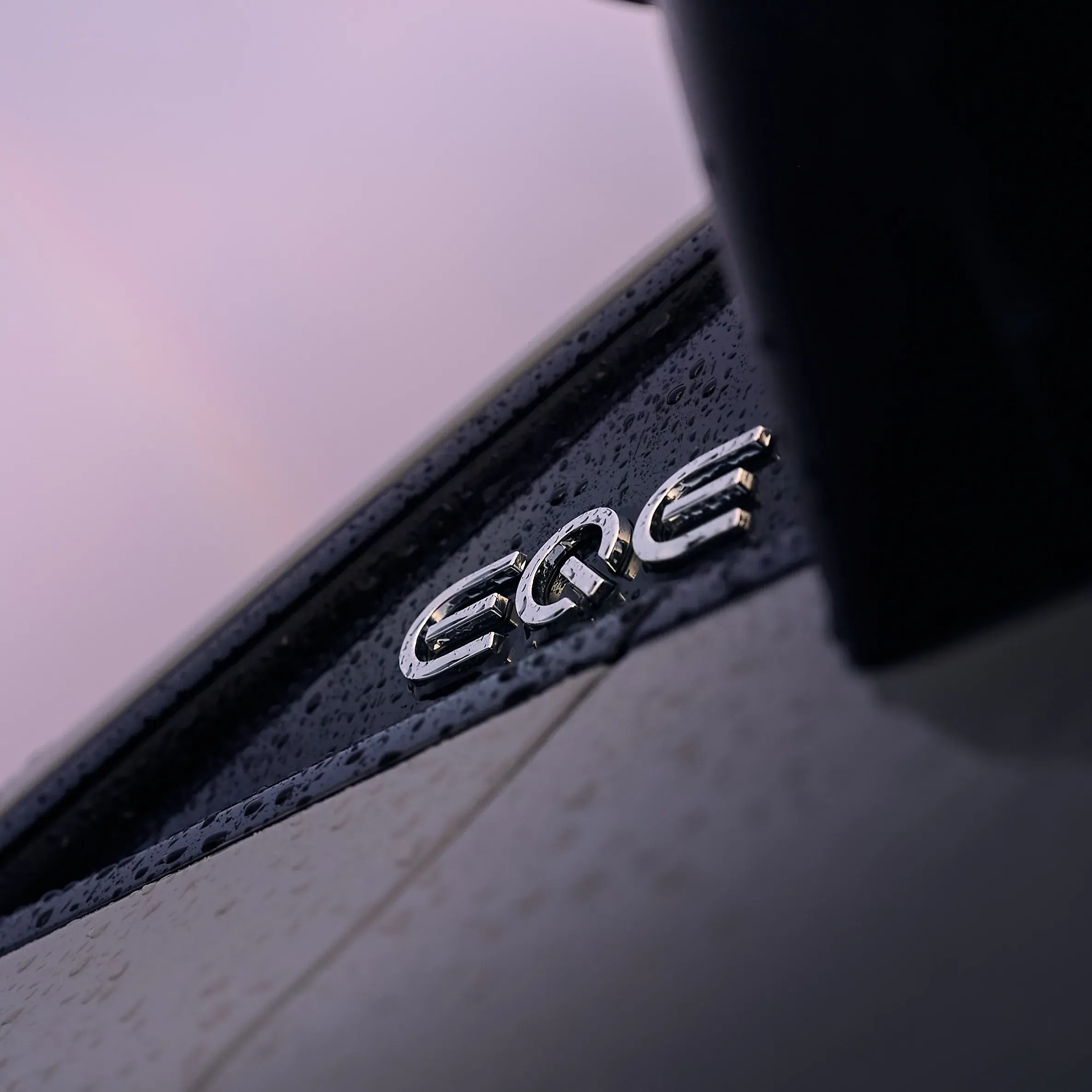 Détail macro d’un monogramme chromé sur carrosserie noire perlée de gouttes de pluie, lumière rose au crépuscule