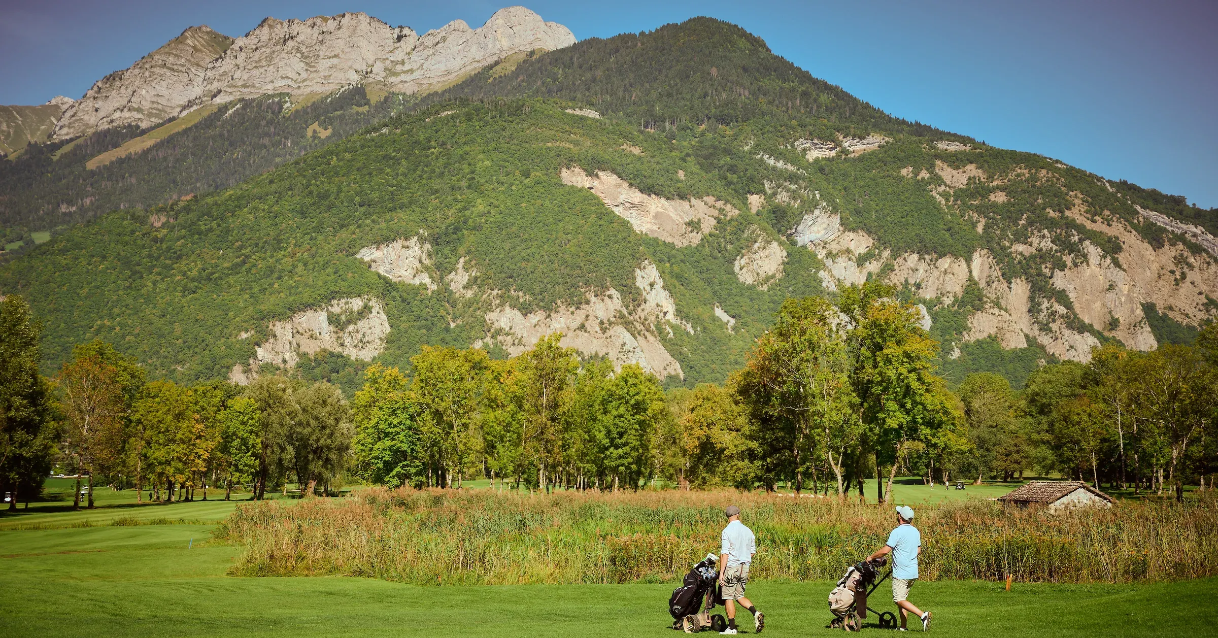 Deux golfeurs tirant leur chariot sur un fairway au pied d'une montagne boisée, à Giez, reportage événementiel