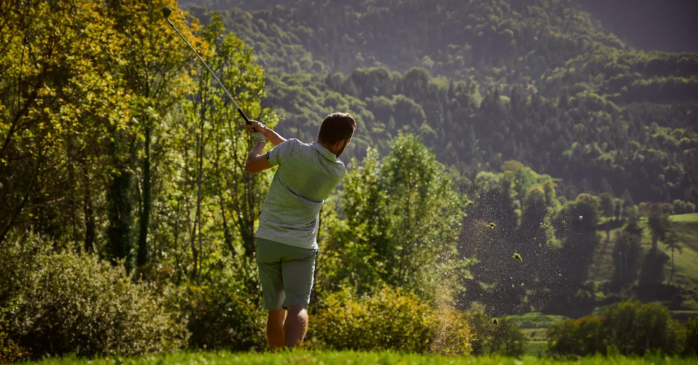 Golfeur en fin de swing, gerbe de gazon dans la lumière, collines boisées en arrière-plan au Golf des Alpes à Giez