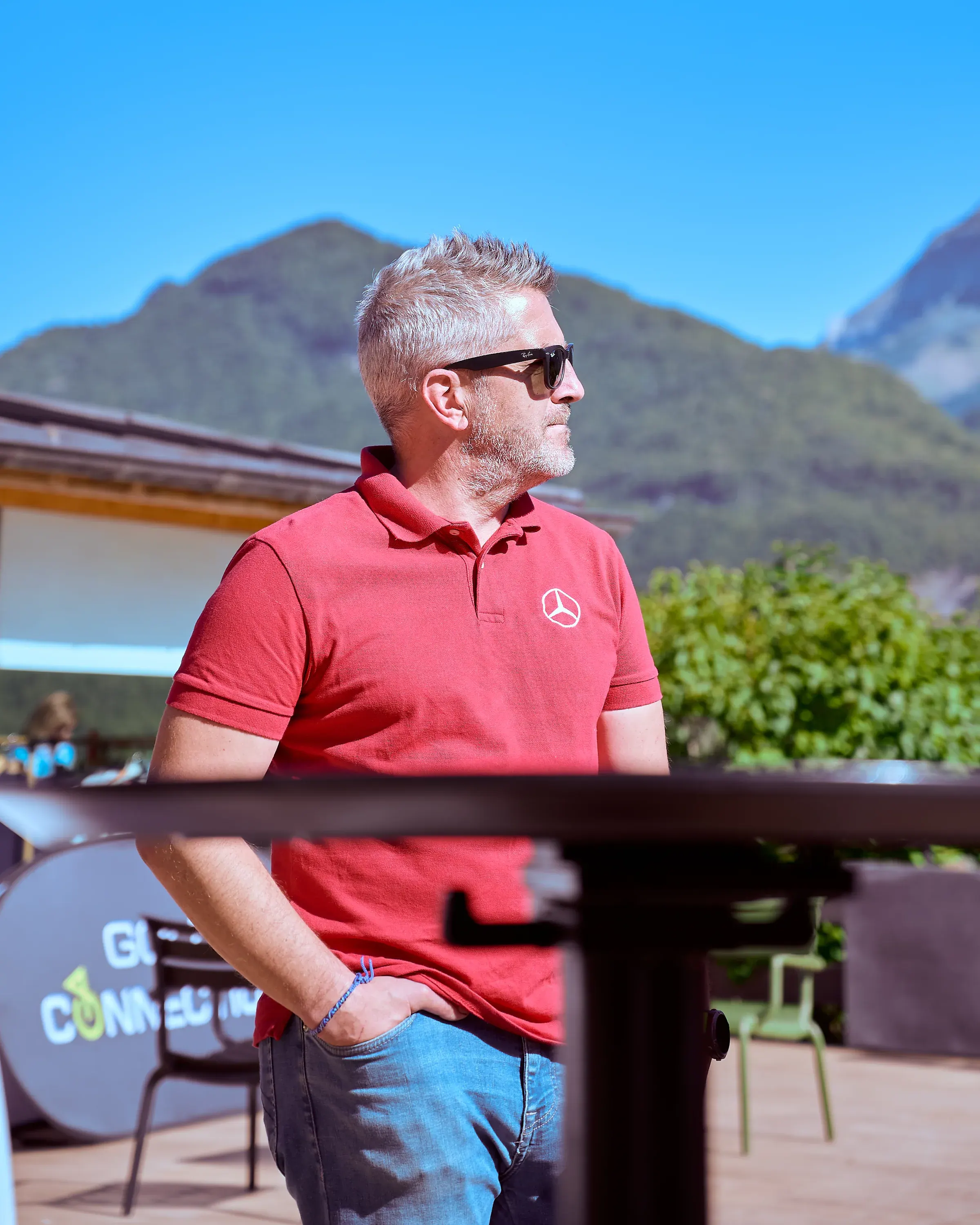 Invité en polo rouge Mercedes-Benz et lunettes de soleil, de profil sur une terrasse face aux montagnes à Giez, reportage événementiel