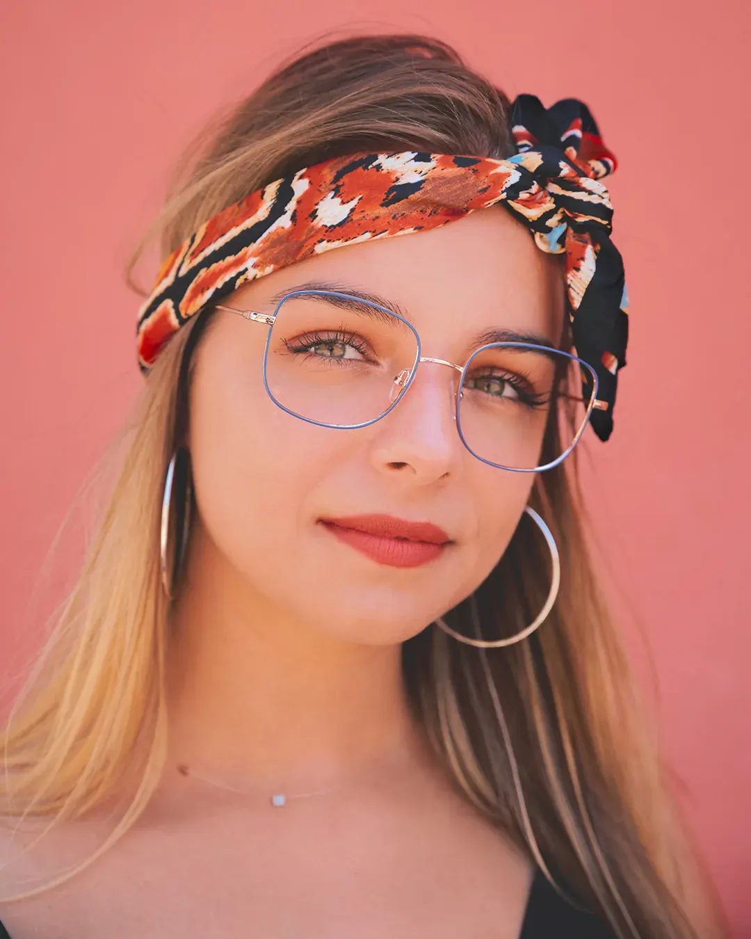 Portrait beauté d’une jeune femme blonde à lunettes et foulard imprimé devant un mur rose, lumière naturelle