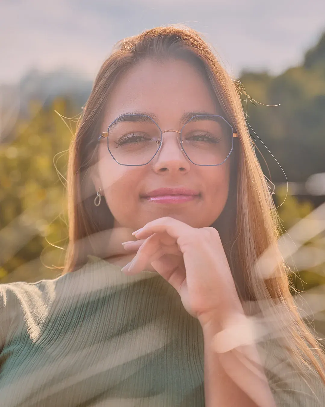 Portrait féminin en extérieur au coucher du soleil, lunettes fines et main sous le menton, lumière dorée et voile de flare