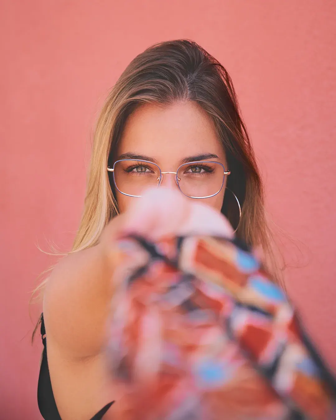 Portrait féminin en lumière naturelle, lunettes fines et regard face caméra devant un mur rose, tissu coloré au premier plan