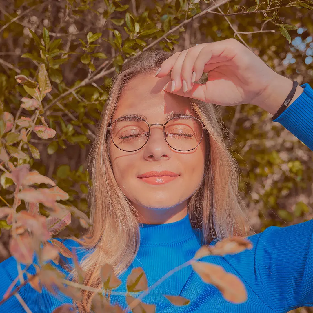 Portrait féminin en lumière naturelle, yeux clos et lunettes rondes, col roulé bleu au milieu du feuillage