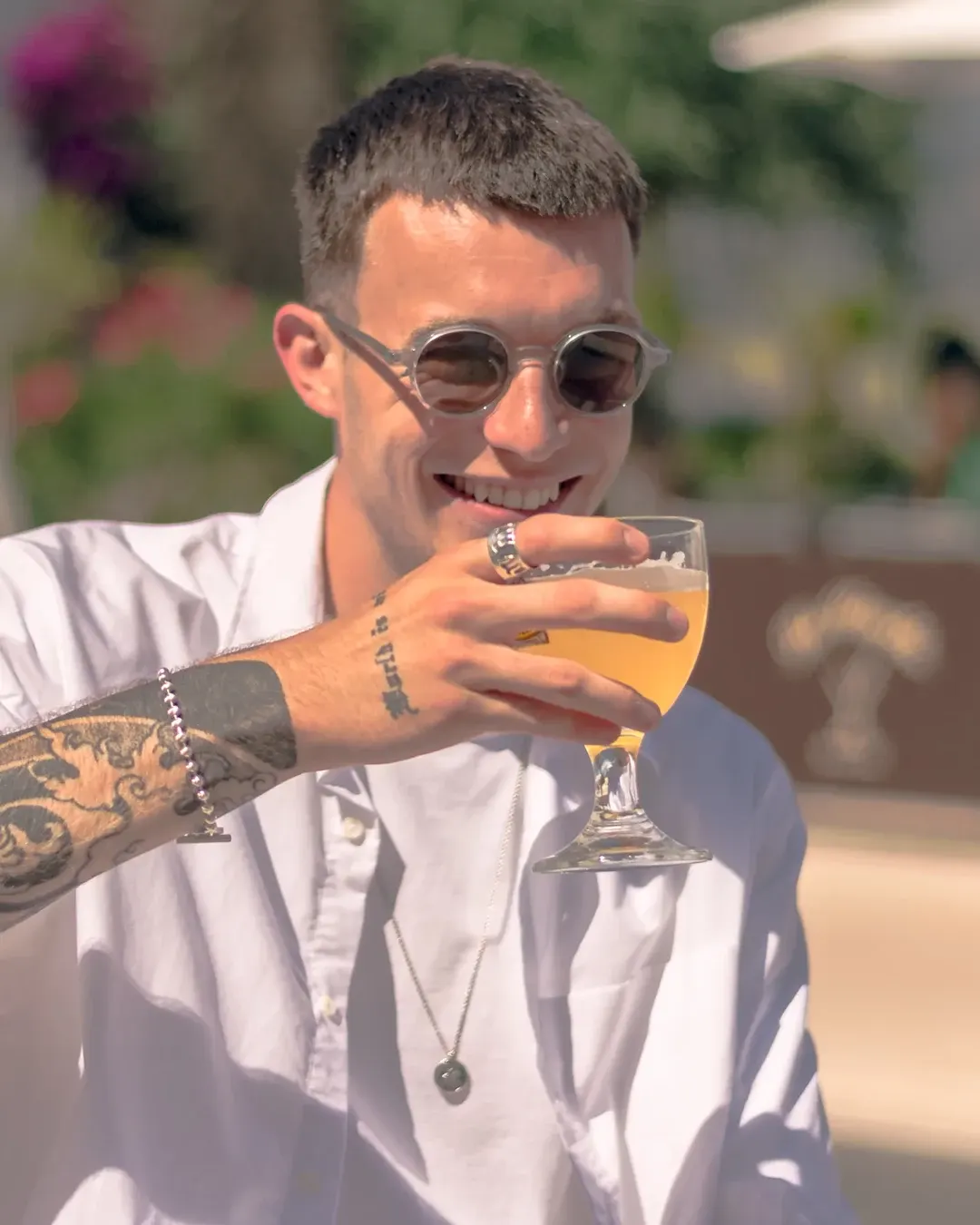 Portrait lifestyle en terrasse : homme tatoué en chemise blanche et lunettes de soleil, souriant, verre à la main