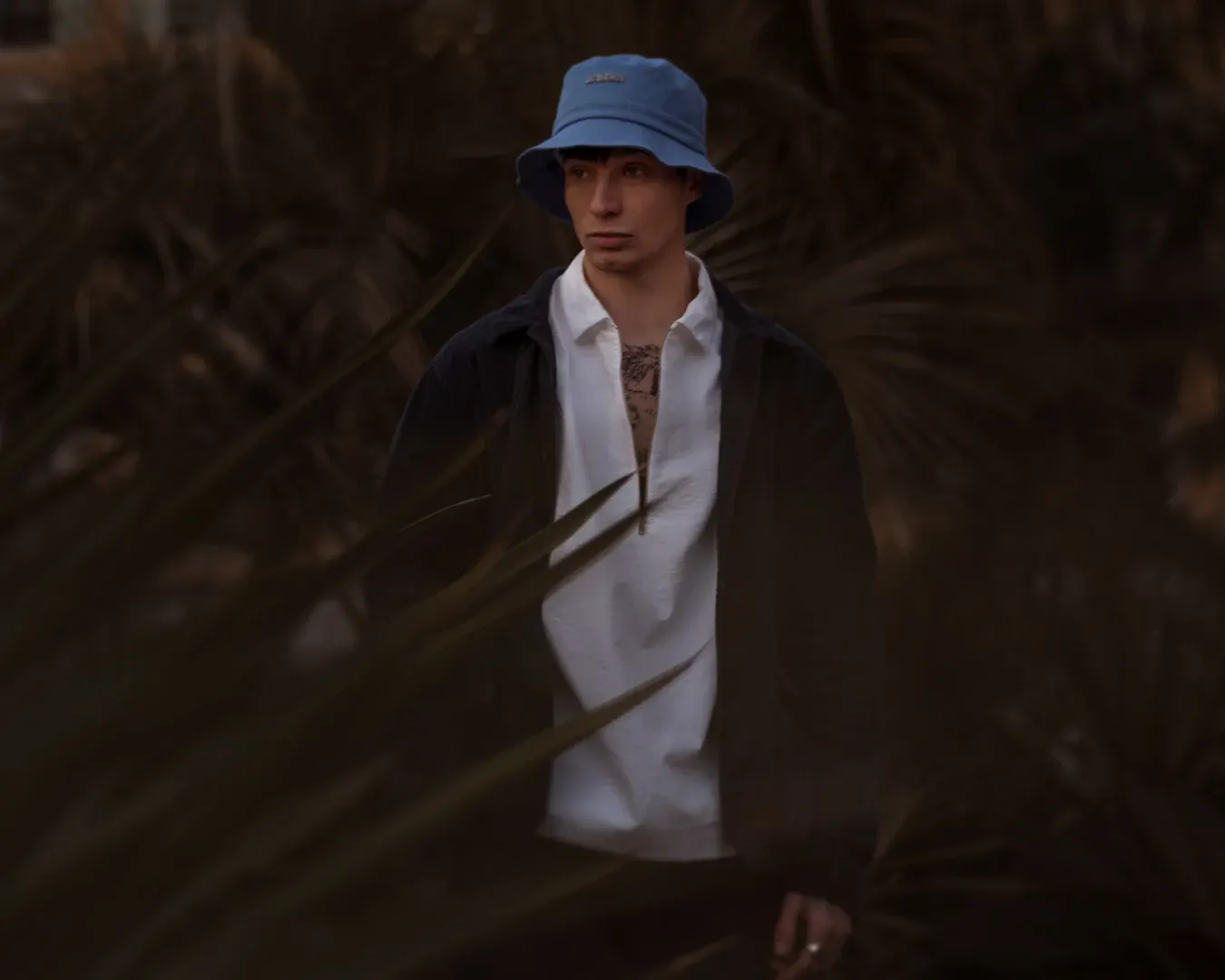 Portrait masculin au bob bleu et polo blanc, cadré à travers des palmes, lumière crépusculaire sur la Côte d’Azur