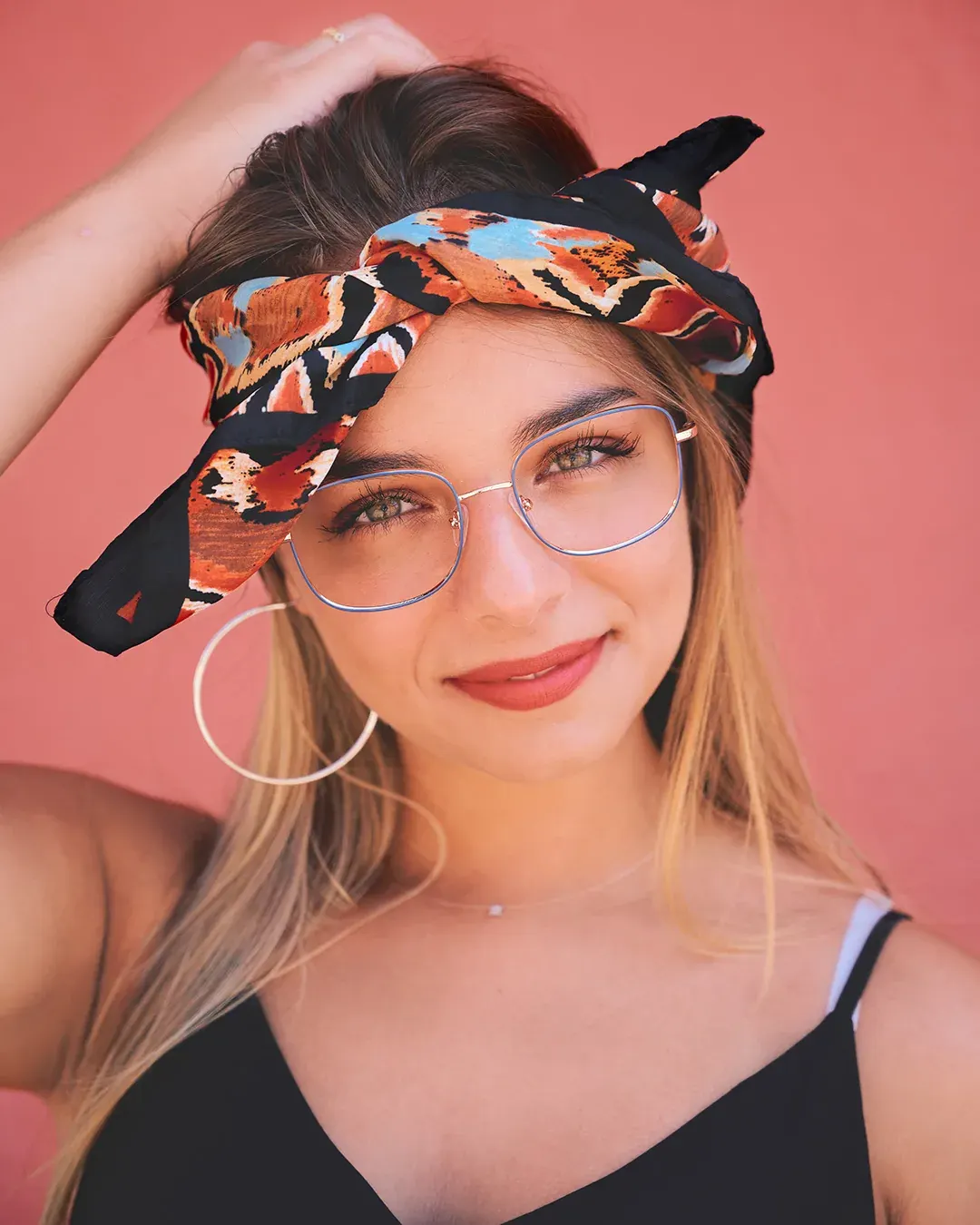 Portrait mode en extérieur : jeune femme au foulard imprimé et lunettes dorées, sourire complice sur mur corail, Côte d’Azur
