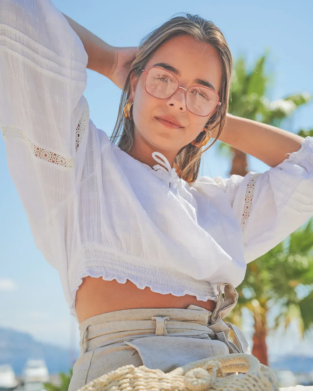 Portrait mode en extérieur d’une jeune femme à lunettes, blouse blanche et palmiers, lumière naturelle sur la Côte d’Azur